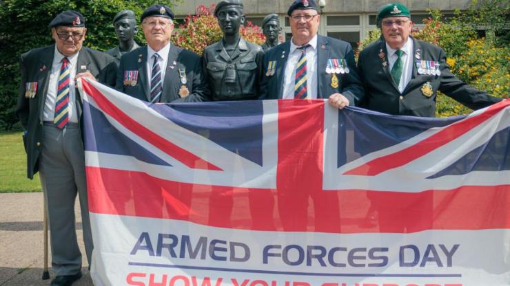 A group of Veterans hold the Armed Forces Day flag, standing in front of a group of statues of soldiers stood at ease.
